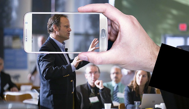 A raised mobile phone capturing a video of a man in a suit giving a public speech