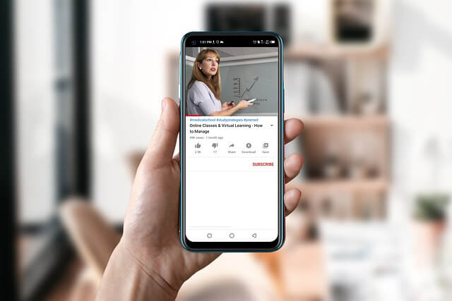 A mobile phone raised in the air with a video playing of a woman teaching a class virtually